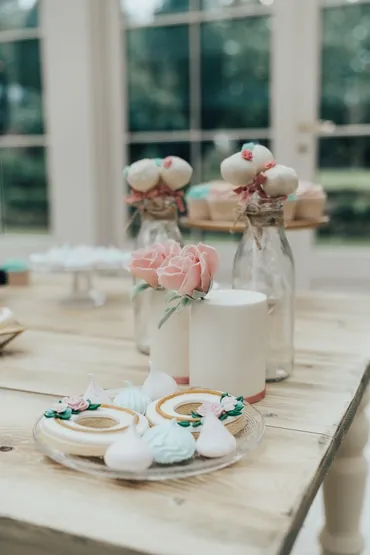 Iced biscuits with meringue kisses on a glass plate alongside cake pops in milk bottles with pink roses on a rustic wooden table