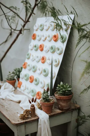 White donut wall display with sage green, orange and white glazed donuts arranged on a board with terracotta pots and greenery