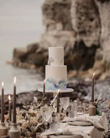 Two-tier white wedding cake with blue watercolour brushstroke detail on a coastal clifftop tablescape