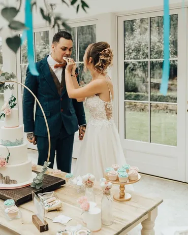 Bride feeding groom wedding cake at a dessert table with cupcakes and teal ribbon decorations