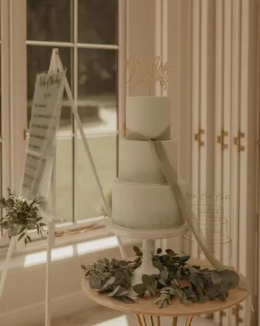 Three-tier white geometric wedding cake with Marry Me Gorgeous gold topper beside a seating plan easel