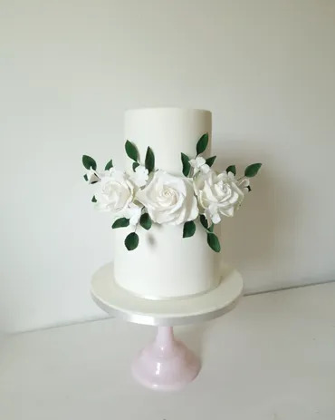 Two-tier white wedding cake with white sugar roses and green leaves on a pink stand