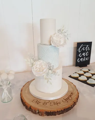 Three-tier white and pale blue geometric wedding cake with white sugar peonies on a Mr & Mrs Spurgeon wooden log stand