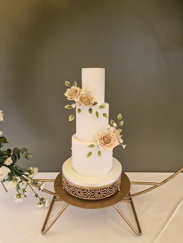 Three-tier white wedding cake with champagne sugar roses and green leaves on a gold stand