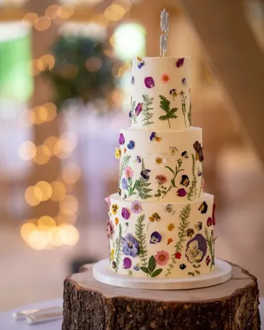 Four-tier white wedding cake covered in pressed flowers and edible botanicals on a wooden log stand