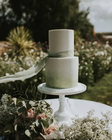 Three-tier wedding cake with pink textured bottom tier, white middle tier and gold drip top tier with vibrant fresh flowers on a wooden log stand