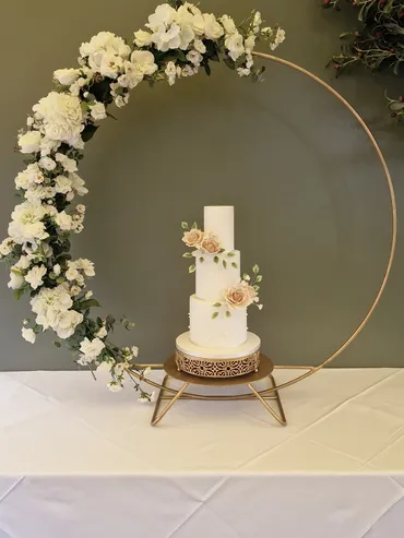 Three-tier white wedding cake with blush roses displayed in a gold hoop arch